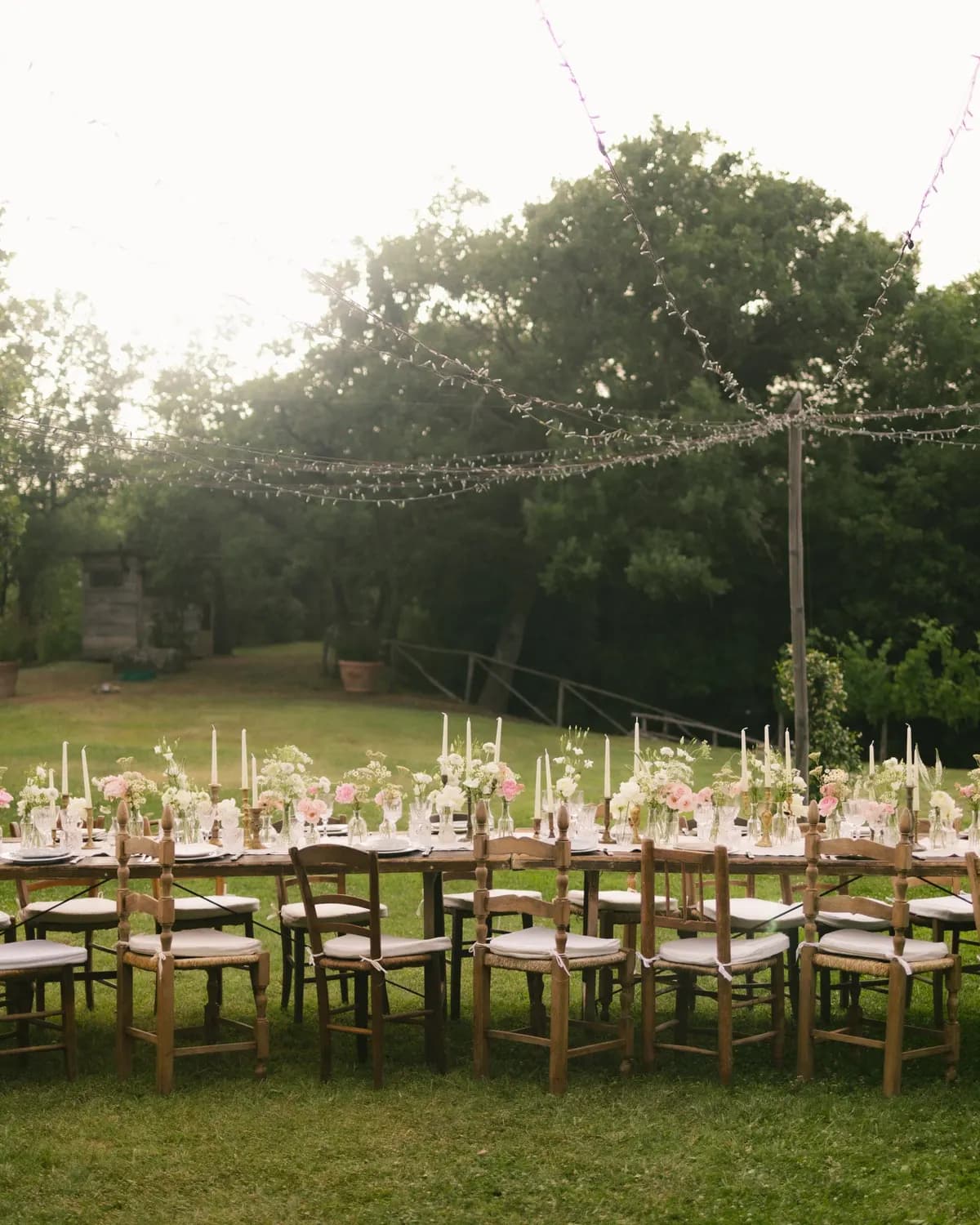 Temporary concept image of long dinner table set on a lawn beneath strings of warm festoon lights; not property photography.